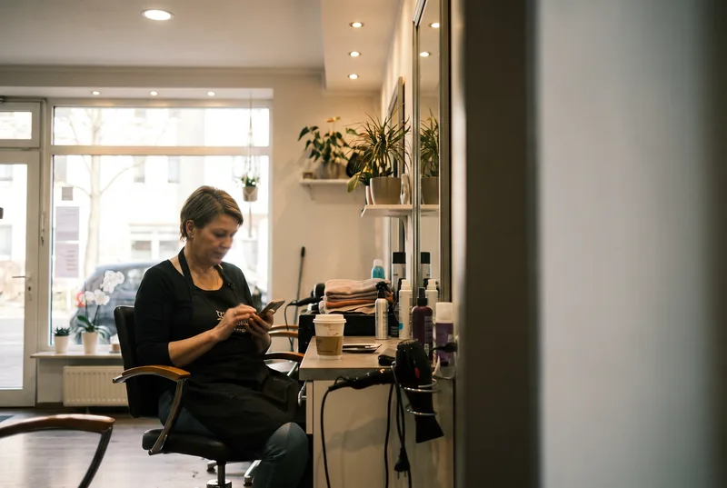 Die Inhaberin eines Friseursalons sitzt morgens vor der Öffnung auf einem Frisierstuhl und scrollt auf dem Telefon durch ihre eigene Webseite